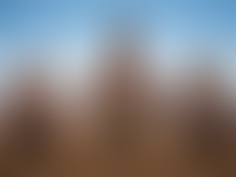 Three men in traditional attire and jewelry stand on brown soil, mountains in the background. They appear solemn under a clear blue sky.