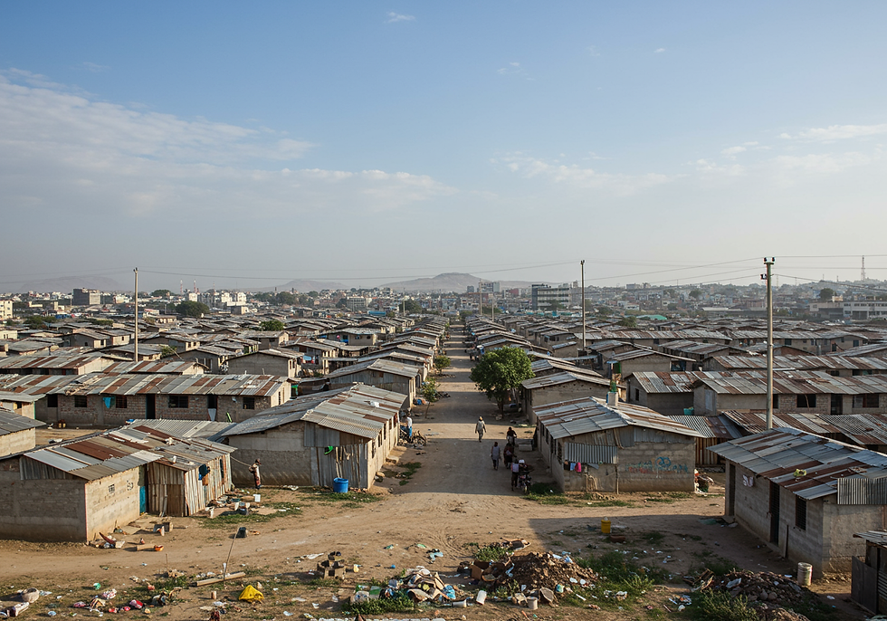 Aerial view of a densely packed settlement with metal-roofed homes. Sparse trees and people walking in paths. Bright blue sky above.