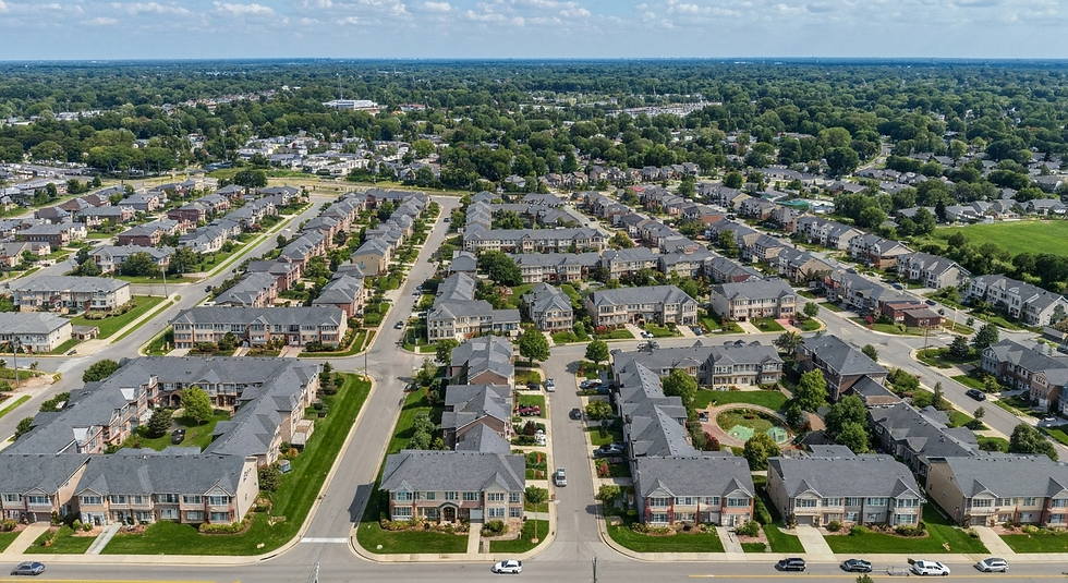 Aerial view of a suburban neighborhood with rows of identical, gray-roofed houses, green lawns, and roads, surrounded by dense trees.