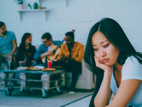 An Asian woman sits away from a group of her peers looking glum. This photo represents the feeling of being unwitnessed or left out.