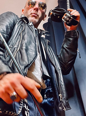 DOM Robert with a leather bomber jacket and sunglasses grips a pair of handcuffs and a whip in style and determination.