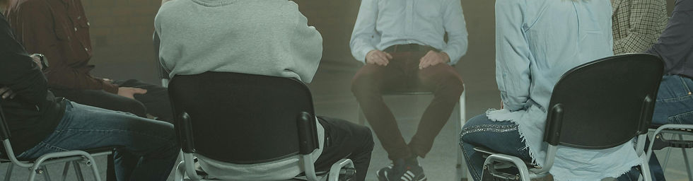 People sitting on chairs in a circle, engaged in discussion. The setting is indoors with a neutral background. Casual attire and calm mood.