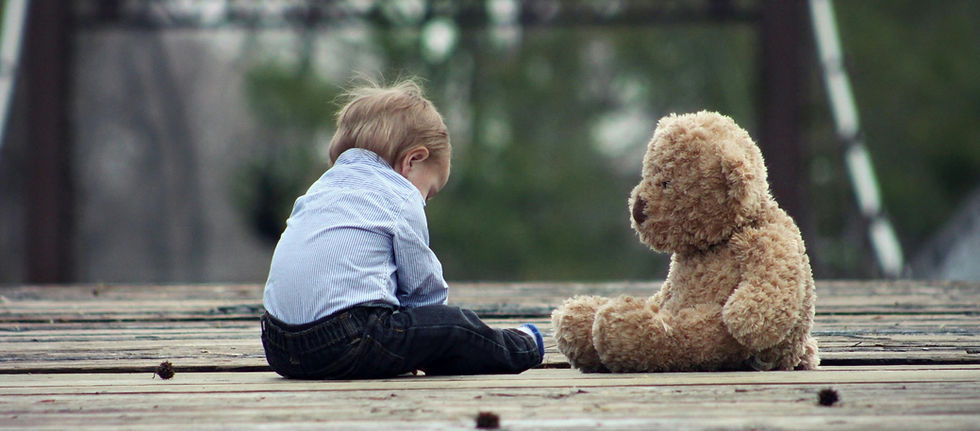 Young child in blue shirt sits on wooden deck facing a fluffy teddy bear. The setting is outdoors with a blurred green background. Mood is calm.