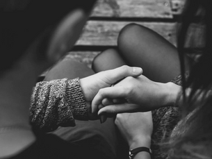 Close-up of two people holding hands on a wooden bench. Black and white image conveys a tender, intimate mood. Sweater texture visible.