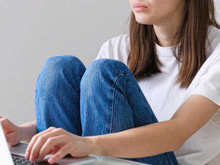Woman in white t-shirt and jeans typing on a laptop at a desk. A glass of water is nearby. Minimalist room with a plant, calm mood.