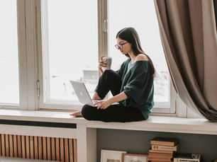 Woman in glasses sits on a windowsill, holding a mug and using a laptop. Sunlight enters through the large window, with a cozy room setting.