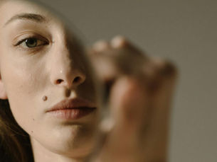 Close-up of a pensive woman with a mole, reflected in a mirror. Soft lighting highlights her features against a neutral background.