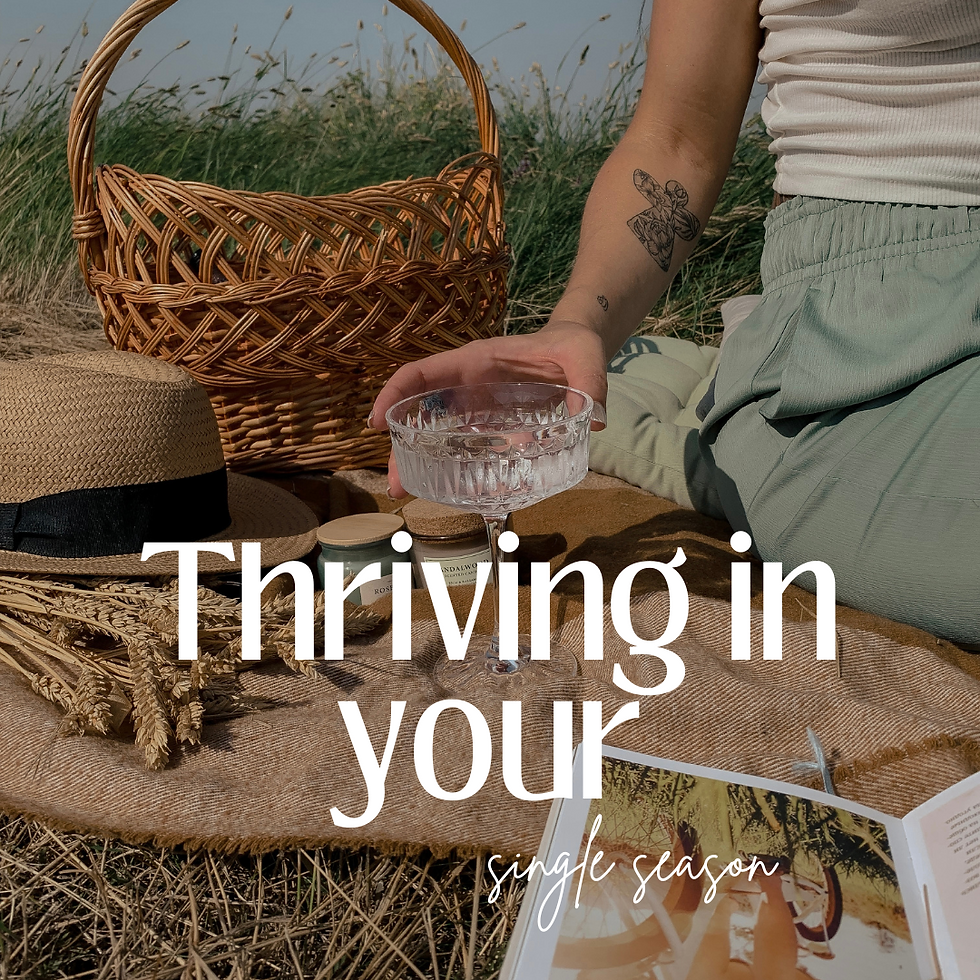 woman sitting on a blanket with a glass in her hand and a picnic basket and hat beside her. tattoo on her arm wearing green pants and white shirt. blue sky behind and tall grass is behind her
