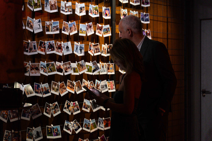 Muro de fotos con personas observando recuerdos: Hombre y mujer viendo una pared llena de fotografías instantáneas en un evento especial.

