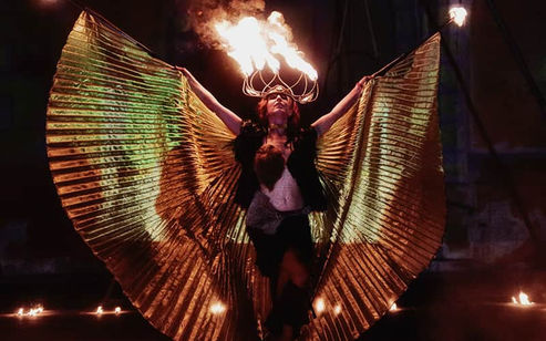 Artista con alas doradas realizando un show de fuego en un escenario oscuro, combinando danza y efectos pirotécnicos.
