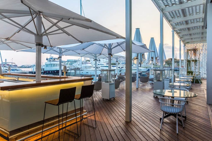 Terraza exterior con sillas altas y sombrillas blancas en zona de bar moderna de madera – bar al aire libre en zona marítima.