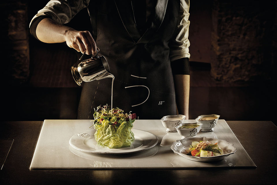 Chef sirviendo una salsa sobre un plato decorado con hojas verdes, con una presentación sofisticada en un entorno elegante.