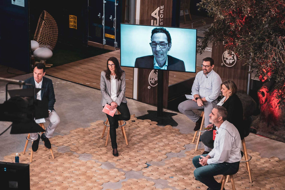 Panel de discusión con participantes en una conversación híbrida, combinando asistentes presenciales y virtuales en un evento empresarial.