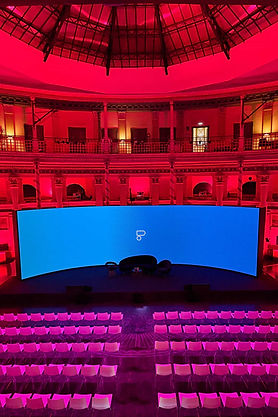 Auditorio en un edificio histórico con arquitectura clásica, iluminación roja y rosa que resalta los detalles. Al frente, un gran escenario curvado con una pantalla azul y filas de sillas blancas perfectamente alineadas.