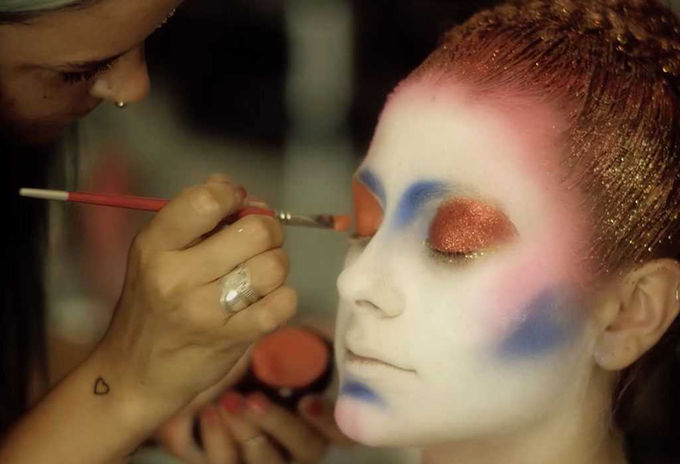 Maquillaje artístico con tonos azules y dorados en el rostro de una mujer – Artista aplicando sombras y detalles creativos en una caracterización teatral.