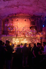 Networking en Integrated Systems Europe (ISE) Barcelona con ambiente industrial y neón – Asistentes socializando en un bar con paredes de ladrillo, iluminación cálida y un letrero de neón "Better Together" en el fondo.