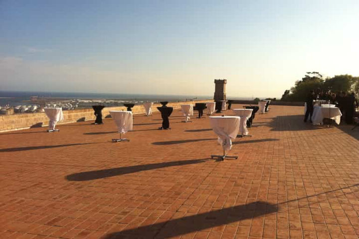 Terraza del castillo preparada para un evento: Mesas altas con manteles blancos y vista al mar al atardecer.
