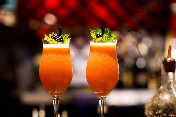 Dos cócteles tropicales de color naranja intenso con decoración de hierbas frescas sobre fondo difuminado – bebidas refrescantes para terraza.