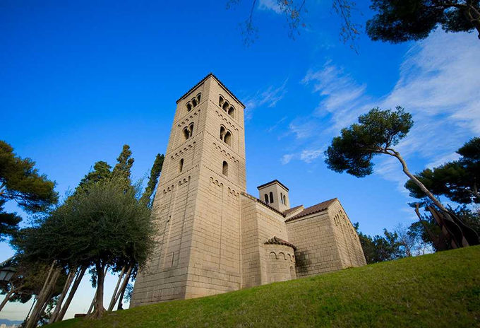 Iglesia románica con campanario en una colina bajo un cielo azul – Edificio histórico rodeado de árboles con arquitectura medieval.