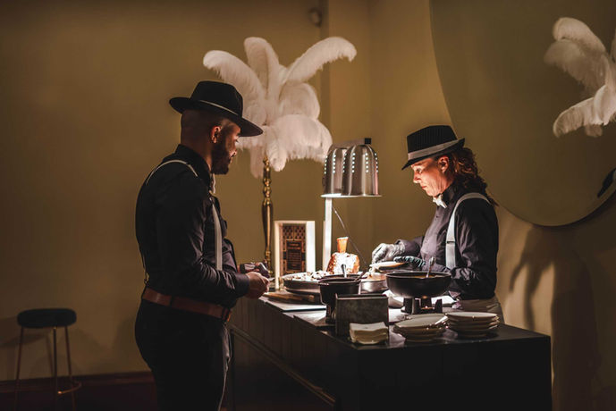 Two people dressed in period themed costumes, serving food at a catering station decorated with white feathers.