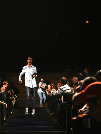 Modelo caminando por una pasarela rodeado de una audiencia que lo fotografía.

