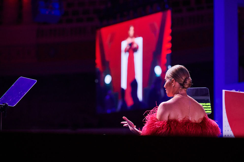 Mujer vestida de rojo en un evento con una gran pantalla detrás, destacando la importancia de la presencia escénica en conferencias y galas.