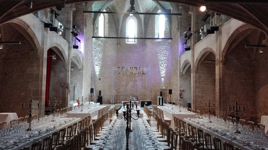 Banquete en iglesia gótica con mesas largas: Cena formal en un amplio salón con techos altos, arcos de piedra y luz natural filtrándose por los ventanales.