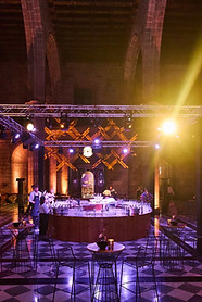 Illuminated bar at a gala event inside a historic building with stone arches – Elegant space with a circular bar, golden lighting and decorated tables in a room with Gothic architecture.