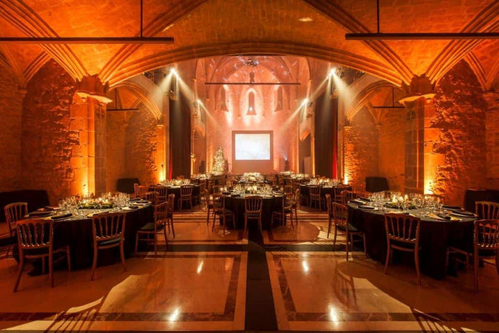 Cena de gala con iluminación dorada en iglesia antigua: Salón decorado con mesas formales y luces cálidas.