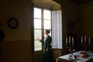 Mujer con atuendo antiguo observando a través de la ventana, reforzando la ambientación de una historia inmersiva.