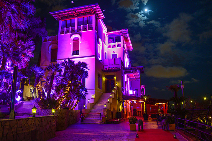 Villa de lujo iluminada en la noche con luces moradas y una gran escalinata rodeada de vegetación tropical.