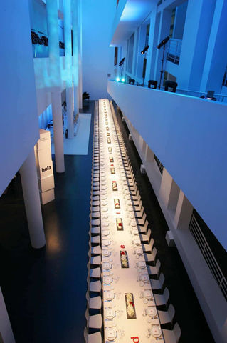 Cena de gala en pasillo iluminado en azul: Mesa alargada con copas y velas en un ambiente sofisticado con luz azul.