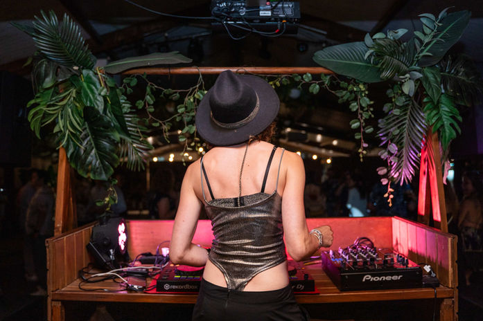 DJ con sombrero negro mezclando música en una cabina decorada con hojas tropicales y luces cálidas.