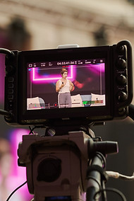 Primer plano de una cámara profesional grabando a una ponente en un escenario, con pantalla LED mostrando contenido digital.
