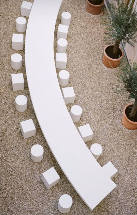Minimalist semicircular table with white chairs and potted plant decorations, designed for an outdoor event.
