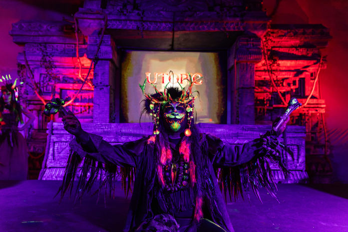 Artista con vestuario tribal futurista y luces de neón en un escenario con el logo "UTOPIC".