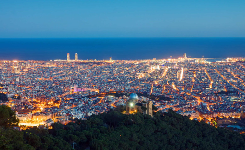 Vista aérea nocturna de Barcelona desde el Observatorio Fabra, con la ciudad iluminada y la cúpula visible en la colina.
