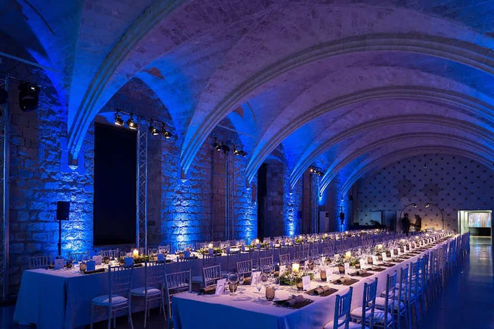 Cena en salón gótico con iluminación azul: Mesas elegantes bajo arcos de piedra con luz tenue en tonos fríos.