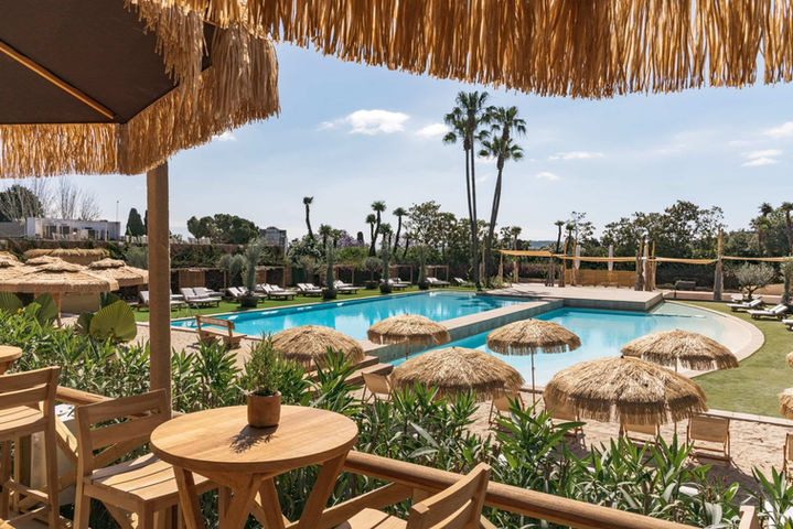 Mesa junto a la piscina con vistas paradisíacas: Pequeña mesa de madera con sombrilla de paja y paisaje tropical.
