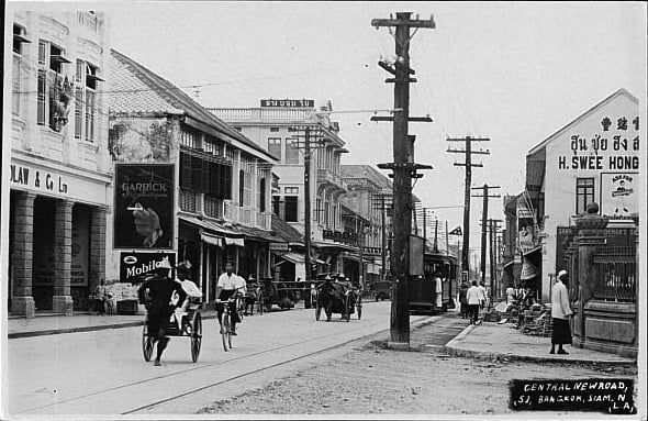 A historical black and white photograph from 1939 showing a bustling street with trams in Bangkok, marking the original Chalern Krung marking time of location of the Kim Seng woodworking shop, the founding lineage of THAVII.