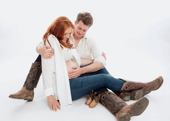 Werdende Eltern auf dem Boden sitzend vor einem weißen Hintergrund in Jeans und Reitstiefeln gekleidet. Bei ihnen stehen noch winzige Reitstiefel in Babygröße - Studioaufnahme aus dem Studio Magic Moments Photographie in Ellscheid nahe Wittlich