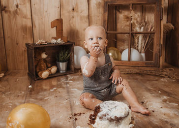 Blondes Kleinkind im grauen Strampler sitzt auf Holzboden mit Kuchenresten um sich herum, schleckt sich genüsslich die Finger beim Cake Smash Shooting zum ersten Geburtstag - liebevoll dekoriertes Set mit Boho - Elementen bei Magic Moments Photographie in Ellscheid, Eifelregion
