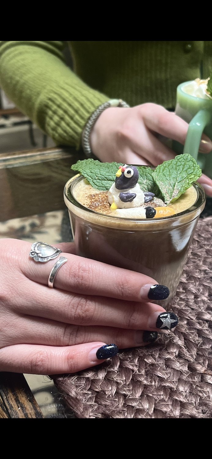 A tea latte topped with a hand-piped meringue character, held in a hand with silver rings.