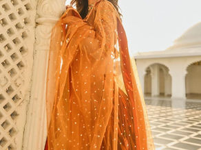 Woman in orange embroidered attire leans against ornate white architecture. Sunlit setting with arches in the background, serene expression.