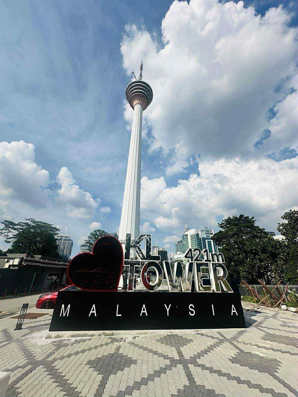 KL Tower stands majestically against a backdrop of fluffy clouds and blue sky, with a vibrant "I ♥ KL Tower" sign welcoming visitors to this iconic landmark in Malaysia.