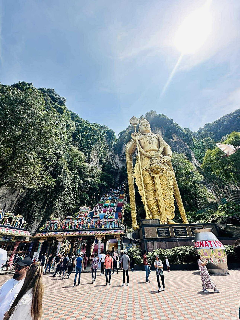 Large golden statue beside colorful steps at Batu Caves. Visitors explore the vibrant temple area. Bright sun and lush greenery in background.