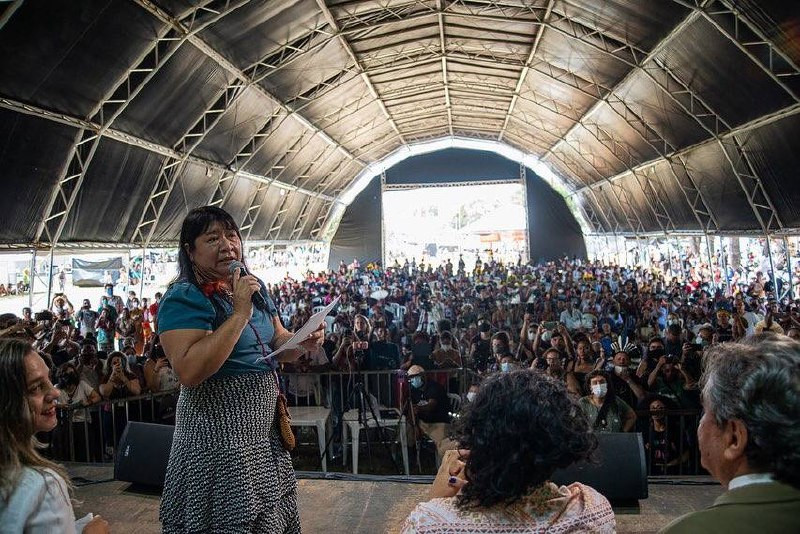 Foto de uma das plenárias do Acampamento Terra Livre. Feita de cima do palco, em primeiro plano mostra a Deputada Federal Joenia Wapichana, em pé enquanto segura um documento e fala ao microfone, de frente para pessoas sentadas no palco que a observam. O que se vê no palco além da deputada são os ombros e cabeças de 3 pessoas, de costas para o fotografo. No plano de fundo está uma numerosa plateia, sob a estrutura de um barracão de metal e lona.