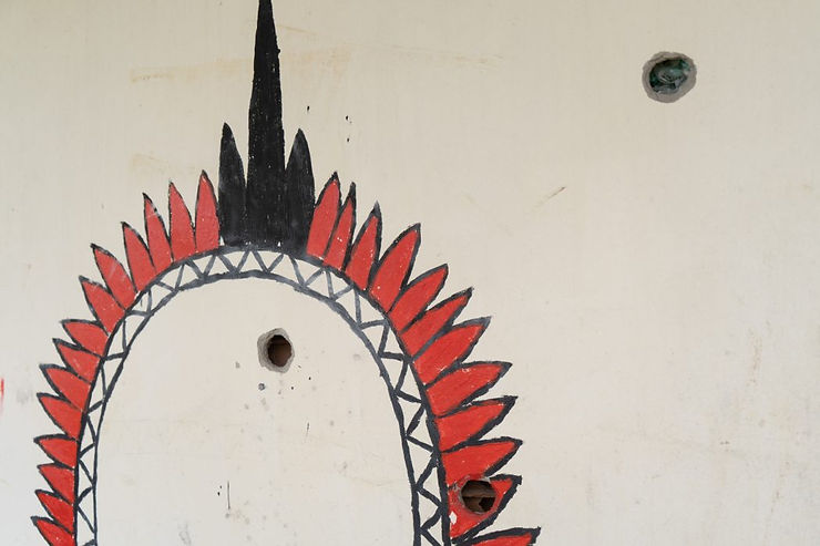 Detalhe de uma parede perfurada com 3 grandes marcas de tiros. Um cocar está desenhado no local das perfurações, penas feitas em tinta vermelha e preta e grafismo em tinta preta.