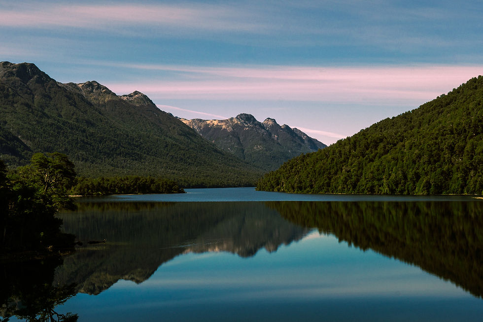 Lago calmo cercado por montanhas e mata, com céu azul e reflexo na água; paisagem serena ao entardecer.