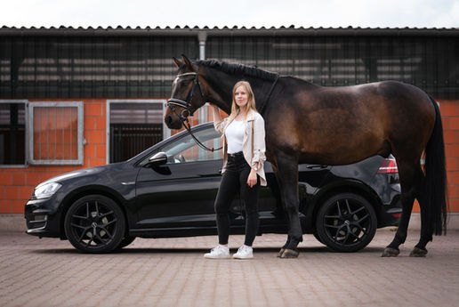 Junges Mädchen steht mit einem großen braunen Pferd vor einem schwarzem VW Golf